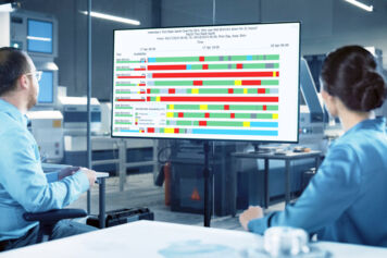 Two equipment engineers in a fab conference room looking at a FabTime Tool State Gantt chart on a monitor.